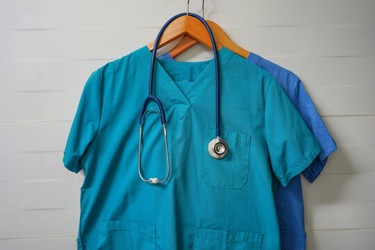 Closeup Doctors Scrus With Stethoscope. Green And Blue Surgical Smock With Stethoscope On Wooden Hanger On Ceramics Wall Background. Closeup Of Doctor's Scrubs With Stethoscope And Lab Coat On Hangers