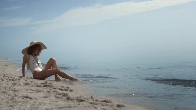 Relaxed woman sitting beach sand washing legs in ocean waves. Girl sunbathing.
