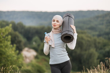 Woman in hijab and sport clothes holding bottle with water while smiling outdoor. Front view of...