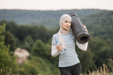 Front view of charming woman in hijab and sport clothes standing at green park and holding yoga mat...