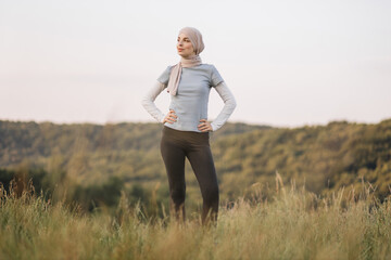 Athletic young Muslim lady in hijab and sportswear standing in sports wear at green summer park, full length. Happy Arabic Islamic woman relaxing after outdoor training.