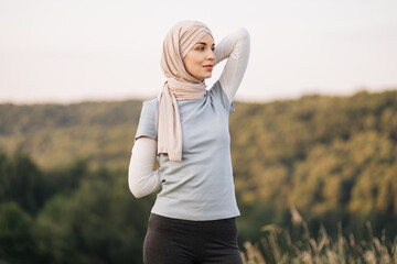 Portrait of sporty muslim woman dressed in religious beige hijab stretching her arms while doing workout in green park outdoors.