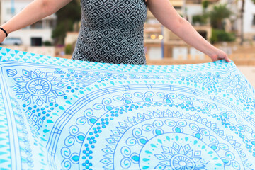 Woman in printed dress spreads blue and white towel on the beach, her face is not visible.