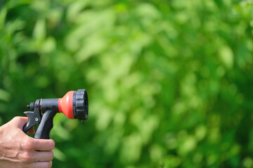 female hand holds a water spray nozzle against the backdrop of a garden and green plants