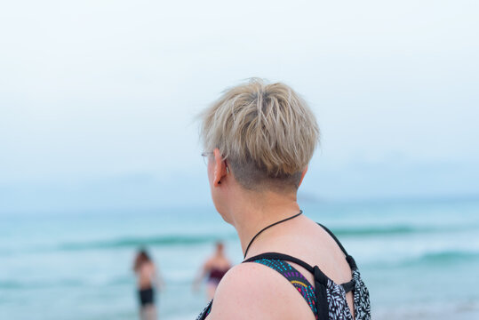 View from behind of a mother watching from the sandy shore her out-of-focus children playing in the water.