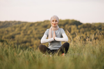 Naklejka na ściany i meble Peaceful young lady in hijab meditating with closed eyes and namaste gesture in the park. Pretty Muslim woman sitting on couch in yoga pose, finding inner balance.