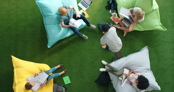 Above view of creative businesspeople planning strategy with paperwork and working on bean bags in informal office setup. Diverse group of excited technology students using fun outdoor startup lounge