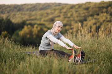 Naklejka na ściany i meble Pleasant woman in hijab and sport clothes sitting on yoga mat and stretching body. Young muslim female training during morning time outdoors.