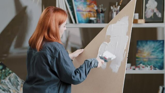 View from behind red-haired Caucasian girl student creative woman artist painter designer scenery picture on canvas cardboard plywood smears white plaster with spatula preparation painting art design