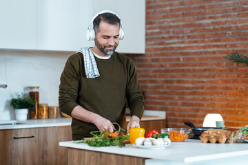 Handsome mature man cutting fresh vegetables while listening music with headphones in the kitchen at home.
