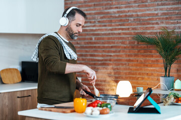 Handsome mature man cooking following a recipe from the internet with digital tablet while...