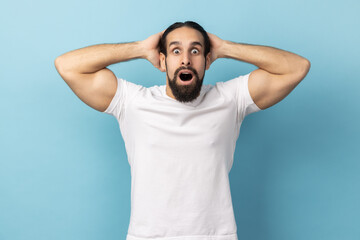 Fototapeta na wymiar Portrait of man with beard wearing white T-shirt standing with mouth open in surprise, has shocked expression, keeping hands on head. Indoor studio shot isolated on blue background.