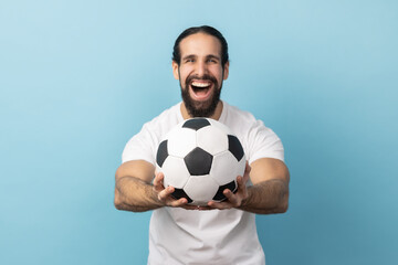 Portrait of man with beard wearing white T-shirt holding out soccer ball in hand and looking at camera with excited facial expression. Indoor studio shot isolated on blue background.