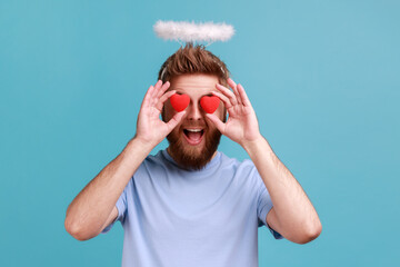Portrait of bearded angelic man with holy nimbus covering eyes with toy hearts as if looking with...