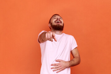 Portrait of funny positive bearded man laughing out loud holding belly and pointing finger on you, mockery, wearing pink T-shirt. Indoor studio shot isolated on orange background.