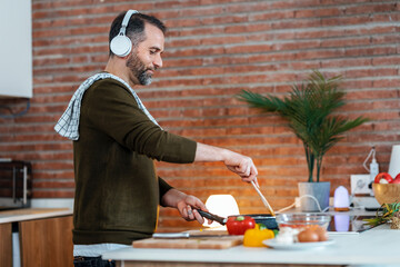 Handsome mature man cooking following a recipe from the internet with digital tablet while listening music with headphones in the kitchen at home.