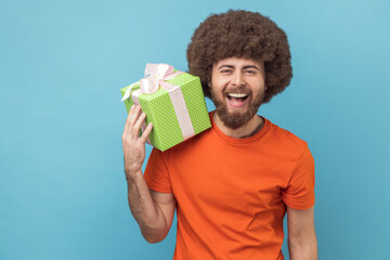 Portrait of extremely happy man with Afro hairstyle wearing orange T-shirt holding green present box and looking at camera with joyful expression. Indoor studio shot isolated on blue background.