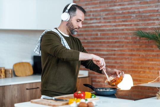 Handsome mature man cooking following a recipe from the internet with digital tablet while listening music with headphones in the kitchen at home.