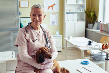 Portrait of smiling senior veterinarian hugging cute dog in vet clinic and looking at camera, copy...