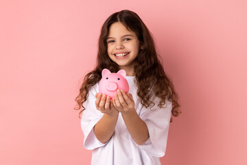 Portrait of little girl wearing white T-shirt holding pink piggy bank, looking at camera with charming smile, best variant for saving money. Indoor studio shot isolated on pink background.