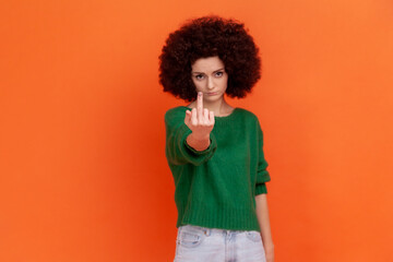 Woman with Afro hairstyle wearing green casual style sweater showing middle finger and asking to get off looking at camera, disrespectful behavior. Indoor studio shot isolated on orange background.