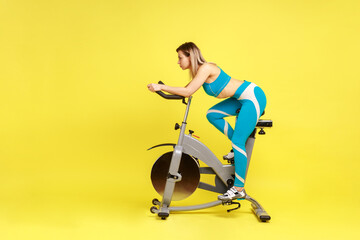 Side view of slim woman with perfect body riding exercise bike at home, making an effort to lose a few extra pounds, wearing blue sportswear. Indoor studio shot isolated on yellow background.