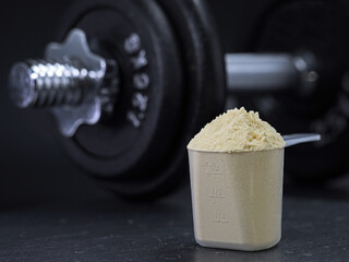 Measuring scoop filled with whey protein powder with dumbbell on black background, protein shake muscle building