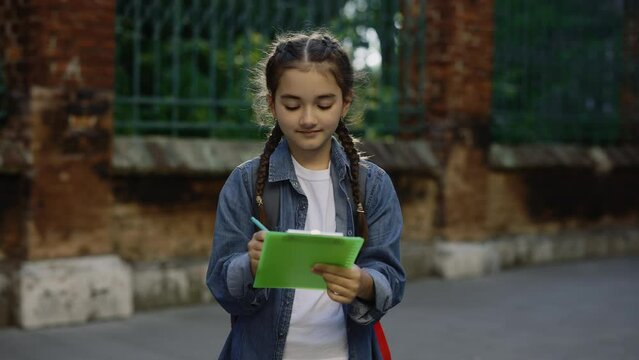 Cute little girl with schoolbag writing notes smiling and looking at camera outdoors slow motion portrait. Portrait. Education concept