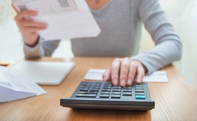 A woman in home clothes is sitting at a table looking through utility bills: gas, electricity, water. counts on a calculator. Selective focus on the calculator.