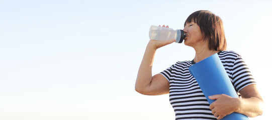 banner of mature woman drinking water from bottle after fitness exercises or yoga outdoors in morning on summer. Fitness female profile on blue sky background, concept of sport and relaxation.