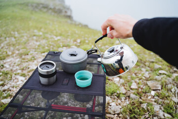 The hand holds the kettle against the background of nature to pour boiling water into a glass on a hike, a tourist on a hiking trip cooks food, brew coffee in the morning, a mountain hike
