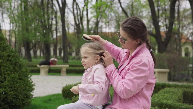 The nanny is combing the girl's hair with a hairbrush while sitting on a bench.