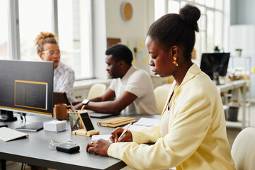 African young woman sitting at table and making notes in her planner with her colleagues working in background