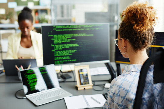 Rear view of young female programmer sitting at her workplace in front of monitors and working with program codes