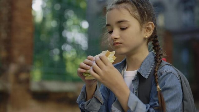A little girl closeup standing on the schoolyard outside looking at food and eating a sandwich with chease, ham and salad. Food and nutrition concept.