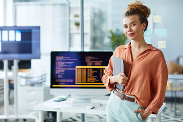 Portrait of young female programmer with digital tablet looking at camera while standing at her workplace with computer
