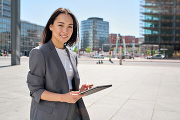 Young happy Asian business woman entrepreneur wearing suit holding digital tablet standing in big...