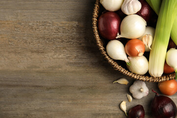 Wicker basket with fresh onion bulbs, leeks and garlic on wooden table, flat lay. Space for text