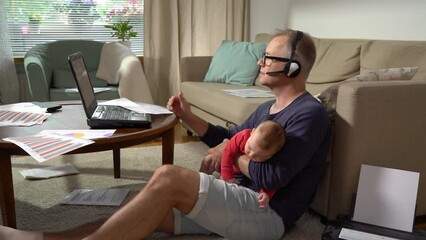Middle-aged father with baby working from home. A freelance single father with newborn makeing a video call and working with documents at home. Remote job, multi-tasking, paternity leave and family.