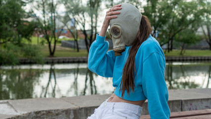A girl in a gas mask sits on a bench on the river bank. Alienation in the city. Fashion, ecology and loneliness concepts.
