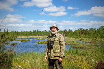 A man with a beard in camouflage clothes, with binoculars, an explorer, a tourist. Stands against the background of a forest lake, forest and blue sky with clouds.