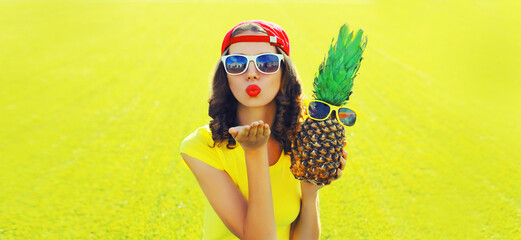 Summer portrait of happy young woman blowing her lips sending air kiss with pineapple and sunglasses on the grass in the summer park