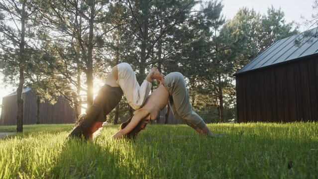 Couple doing acro yoga in yard. Zoom out and in view of man and woman bending forward and doing acro yoga on grass against trees and sunrise sky in morning in yard
