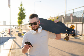 Young man holding on his longboard and using a smartphone while walking in the city.
