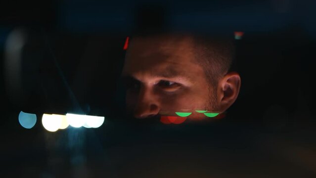 Close Up Of Young Serious Man Inside The Car In Evening. Tired Caucasian Male Driving His Car And Looking At Rear View Mirror. Blurred Background. Night Time. Traveling Concept