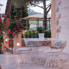 Gorgeous corner of romantic terrace with white vintage metal table and chairs on a stone tile floor. Stone building and trees in the background and deep pink flowers in the foreground.
