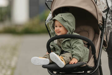 A happy female toddler is sitting in the stroller on a cloudy day. In a green village, a young girl...