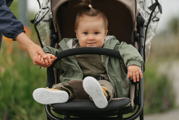 A female toddler is sitting in the stroller on a cloudy day. In a green village, a young pretty girl in a raincoat is in a baby carriage. A mother is holding the hand of her daughter.