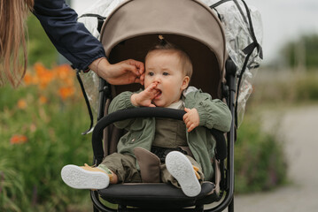 A female toddler is putting her fingers in her mouth in the stroller on a cloudy day. In a green village, a young pretty girl in a raincoat is in her baby carriage.