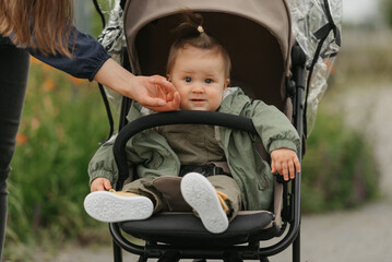 A female toddler is sitting in the stroller on a cloudy day. In a green village, a young pretty girl in a raincoat is in a baby carriage. A mother is touching the cheek of her daughter.